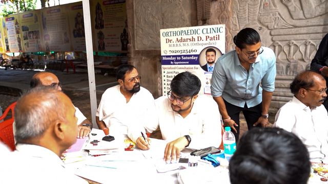 A standing signboard for the clinic beside the consultation table, with Dr. Adarsh Patil seated with attendees during the camp
